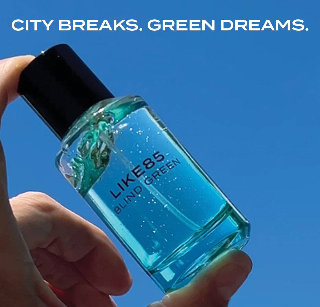 Hand holding a blue bottle of LIKE85 Blind Green perfume against a clear blue sky with text 'CITY BREAKS. GREEN DREAMS.'